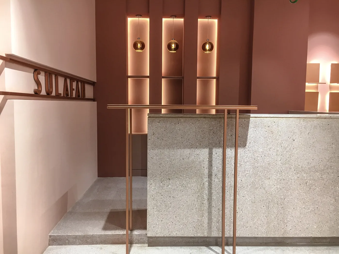 Terrazzo reception counter at Sulafah, lit against terracotta walls and copper shelving