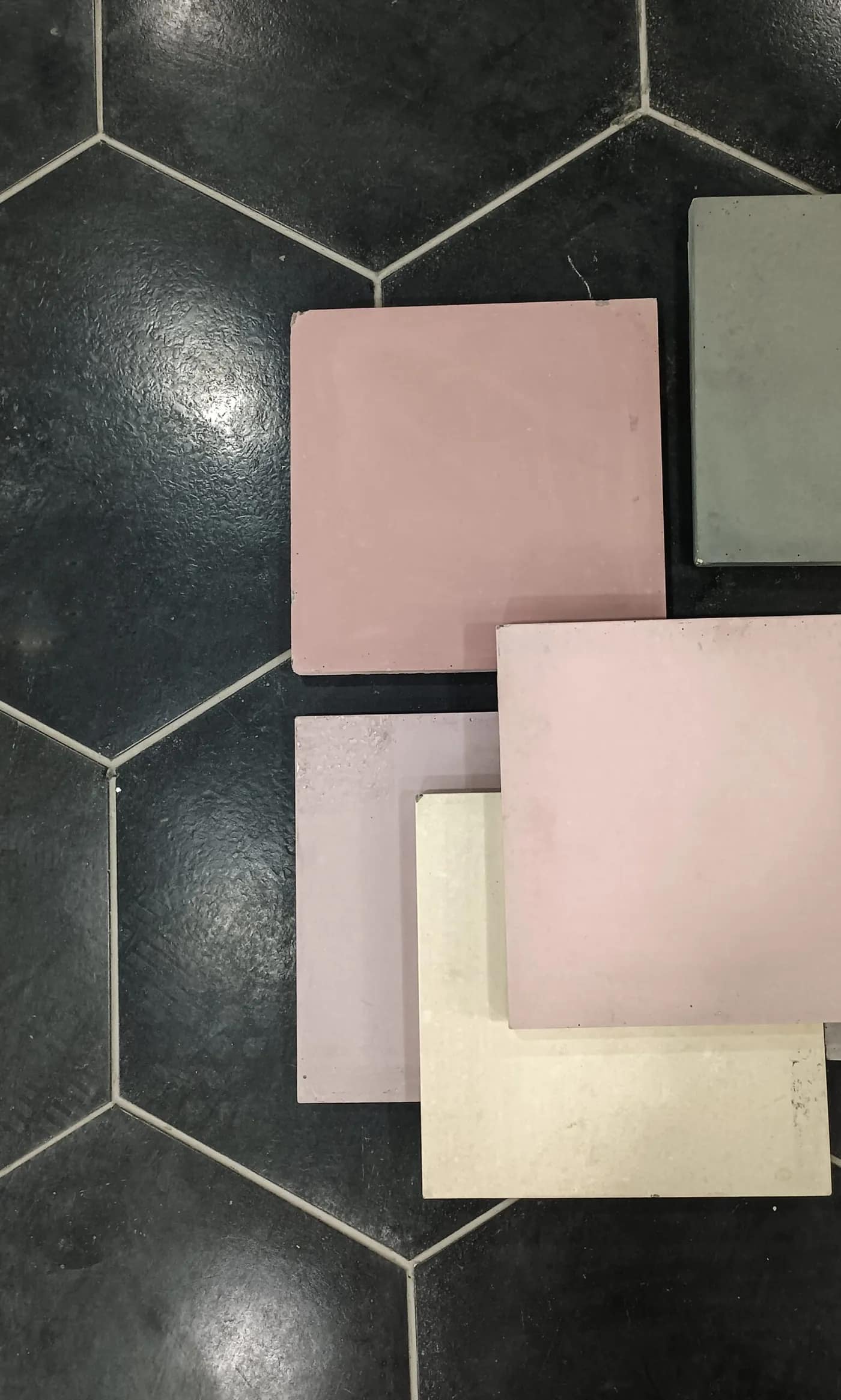 Five plain pigmented cement tiles in dusty pink, mauve, sage and cream, laid loose on a dark hexagonal tiled floor